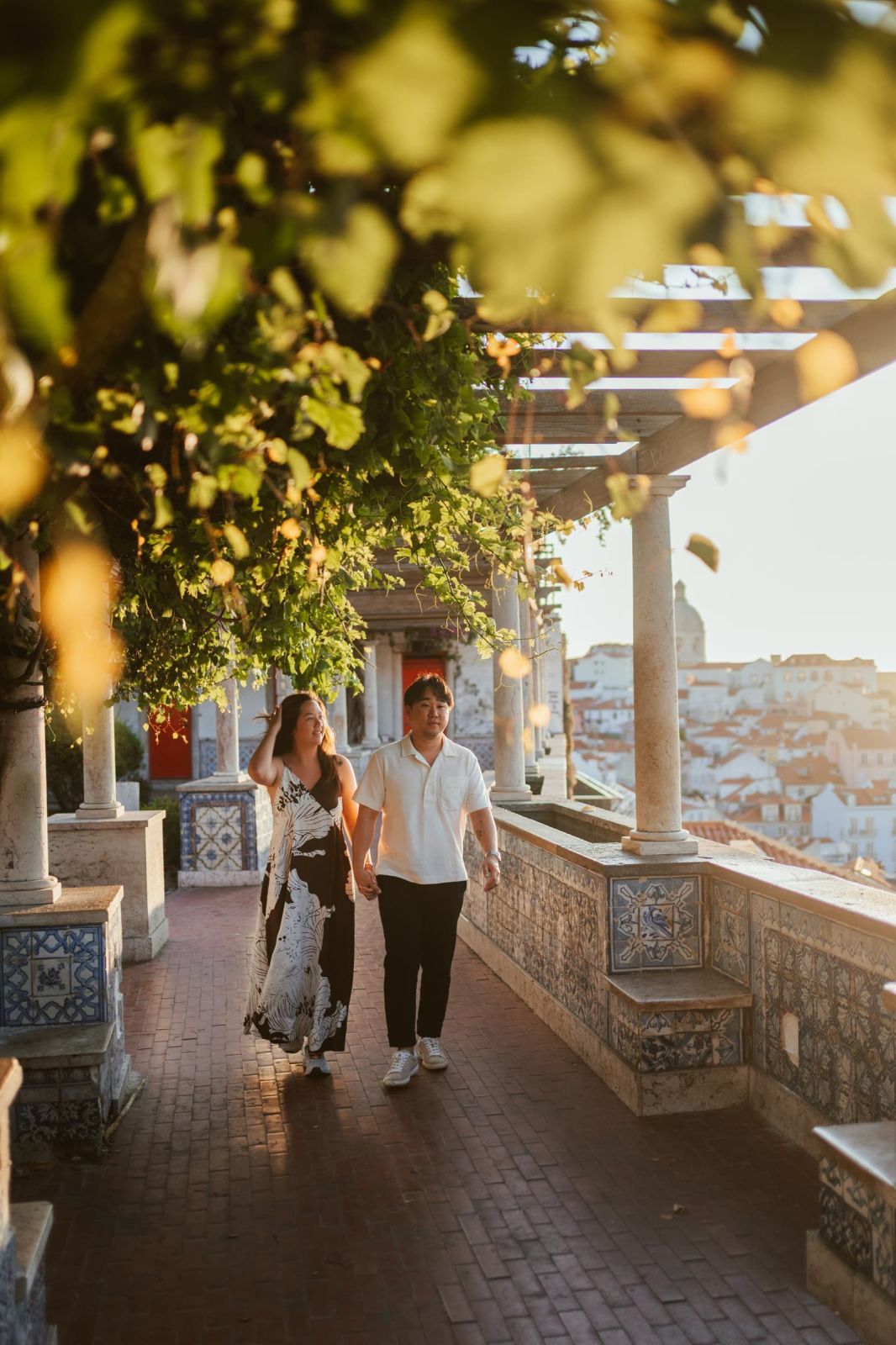 Couple walking under a vine-covered pergola at a Lisbon viewpoint over Alfama